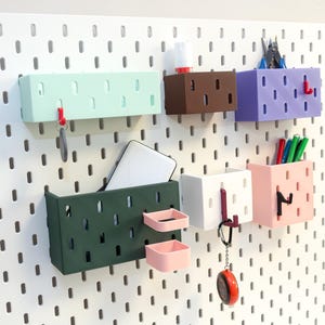 Peut inclure: Ensemble de boîtes de rangement murales colorées de différentes formes et tailles. Les contenants sont dans des tons de vert, marron, violet, blanc et rose. Certains contiennent des clés, des stylos et un téléphone. Montés sur un panneau perforé blanc.