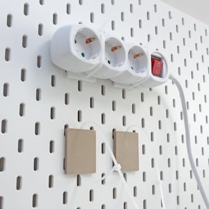 Peut inclure: Une multiprise blanche avec trois prises électriques et un interrupteur rouge lumineux fixée sur un panneau perforé blanc. Deux petites plaques de montage carrées de couleur beige clair sont fixées au panneau sous la multiprise avec des câbles.