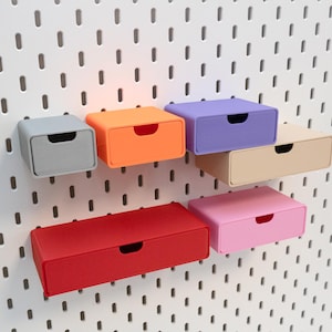 Op de afbeelding: Zes kleine, kleurrijke opberglades in grijs, oranje, paars, beige, rood en roze zijn gemonteerd op een wit pegboard. Elke lade heeft een kleine, gebogen handgreep. De laden zijn rechthoekig en lijken van plastic te zijn.
