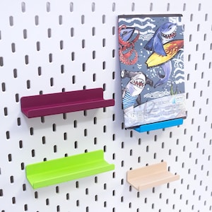 May include: A white pegboard with four colorful shelves and an exercise book. The shelves are magenta, green, and beige. The exercise book has a shark illustration and the text "EXERCISE BOOK".