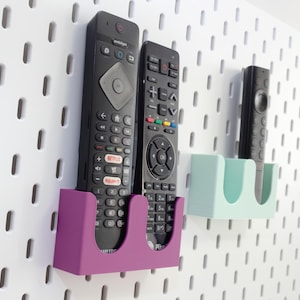 Può includere: Una pannello forato bianco con tre telecomandi in supporti. I supporti sono viola e azzurro. I telecomandi sono neri con vari pulsanti. Il pannello forato ha fori ovali distanziati.