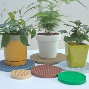 Puede incluir: Seis posavasos redondos y texturizados de diferentes colores, incluyendo azul, marrón, beige y verde. Los posavasos están dispuestos sobre una superficie blanca. Se pueden ver tres plantas en macetas en el fondo.