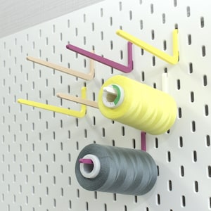 May include: A white pegboard with yellow, pink, and beige hooks holding spools of yellow and gray thread.