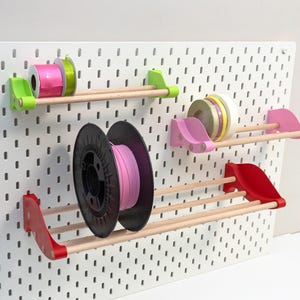 Könnte beinhalten: Eine weiße Lochwand mit Holzregalen, die Spulen mit Bändern und Filamenten halten. Die Regale werden von farbigen Halterungen in Grün, Rosa und Rot getragen. Die Spulen sind in verschiedenen Farben erhältlich, darunter Rosa, Grün und Weiß.