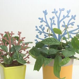 Puede incluir: Dos plantas en maceta con siluetas de árboles metálicas decorativas. La planta de la izquierda tiene ramas marrones y hojas verdes. La planta de la derecha tiene hojas verdes y una silueta de árbol metálica azul.