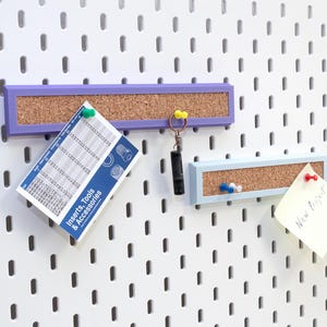 Pode incluir: Um painel perfurado branco com duas placas de cortiça retangulares, uma roxa e outra azul clara. Um cartão com o texto "Inserts, Tools & Accessories" está preso ao painel. Um chaveiro e um post-it também estão anexados.