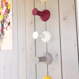 Peut inclure: Cinq patères murales colorées, crème, bordeaux, blanche, grise et jaune, sont fixées sur un mur en panneaux de bois clair. Les crochets ont une base arrondie et une projection cylindrique, offrant un design moderne.