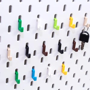 May include: A white pegboard with various colored hooks and a set of keys hanging from one. The hooks are in shades of green, blue, yellow, black, brown, and beige. The pegboard has evenly spaced oval holes.