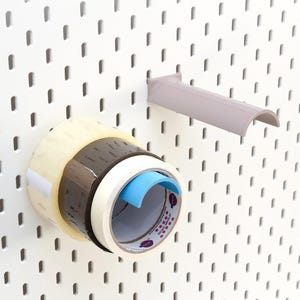 Peut inclure: Un panneau perforé blanc avec des rouleaux de ruban adhésif et un distributeur de ruban gris. Les rouleaux de ruban sont transparents, marron et blancs, avec un coupe-ruban bleu. Le distributeur est monté sur le panneau.