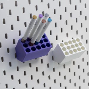 May include: A purple and a white rectangular plastic pen holder, each featuring twelve circular holes for storage. The purple holder contains four markers with the brand name Cricut printed on the barrels. Both holders are mounted on a white pegboard.