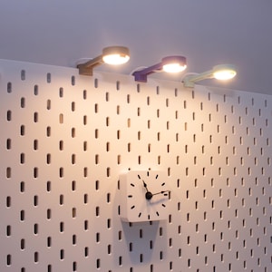 May include: Three small, colorful LED lamps are mounted on a white pegboard. The lamps are in shades of beige, purple, and light green. A white square clock with black hands and markers is also mounted on the pegboard.