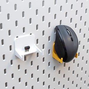 May include: A white pegboard with a black computer mouse and a USB receiver, each held by a 3D-printed holder. The mouse holder is yellow, and the USB receiver holder is white. The pegboard has evenly spaced holes.