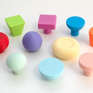 Może przedstawiać: Kolekcja kolorowych gałek do szafek w różnych kształtach, w tym okrągłych, kwadratowych i stożkowych. Gałki są w odcieniach czerwieni, zieleni, fioletu, niebieskiego, żółtego, pomarańczowego i różowego. Gałki wykonane są z teksturowanego materiału.