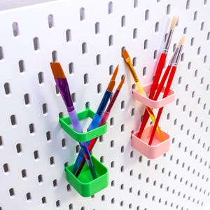 Puede incluir: Un tablero perforado blanco con dos juegos de pinceles coloridos en soportes verdes y rosas. Los pinceles tienen mangos y cerdas rojas, moradas, azules y amarillas.
