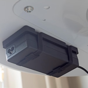 May include: A black power supply unit is mounted under a white surface, secured with a black strap. The unit has an AC power inlet and a black cable extending downwards. The background is a white surface with a few screws.