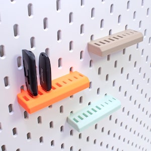 Può includere: Una lavagna forata bianca con diversi ripiani colorati e due chiavette USB nere. I ripiani sono arancioni, beige e verde menta. La lavagna forata ha fori distanziati per il montaggio dei ripiani.