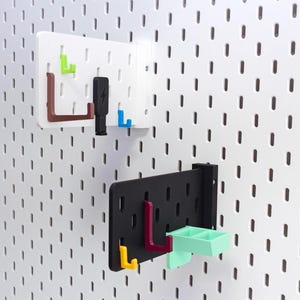 Puede incluir: Dos organizadores de panel perforado con varios ganchos y accesorios de colores. Uno es blanco, el otro negro. Los ganchos son verdes, marrones, azules, amarillos y rojos. También se incluye una pequeña bandeja verde menta. Los paneles perforados están montados en una pared blanca.