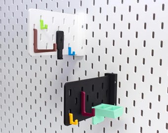 May include: Two pegboard organizers with various colored hooks and accessories. One is white, the other black. The hooks are green, brown, blue, yellow, and red. A small mint green tray is also included. The pegboards are mounted on a white wall.