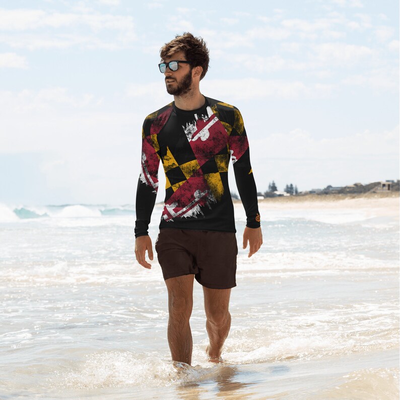 May include: A man wearing a black long-sleeve shirt with a distressed Maryland flag design. He is standing on a beach with the ocean in the background.