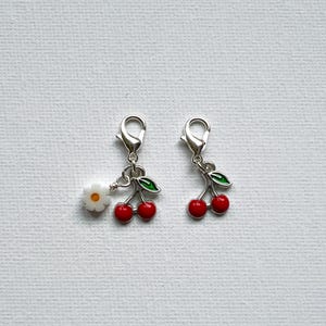 May include: Two silver-tone charms with lobster clasps. One charm features a white daisy, the other has a pair of red cherries with green leaves. These charms are ideal for jewelry making or adding to a keychain.
