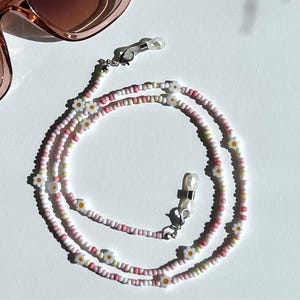 May include: A beaded eyeglass chain with a daisy flower design. The chain is made of small pink, white, and yellow beads. The chain is connected to the glasses with silver-colored loops. A pair of sunglasses is in the upper left corner.