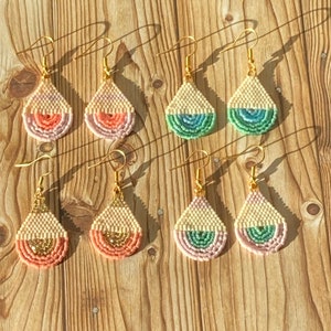 May include: A selection of handmade teardrop earrings with gold-coloured hooks. The earrings have a beaded design with a cream-coloured top and a colourful, semi-circular bottom in shades of pink, coral, green, and blue. The earrings are displayed on a wooden surface.