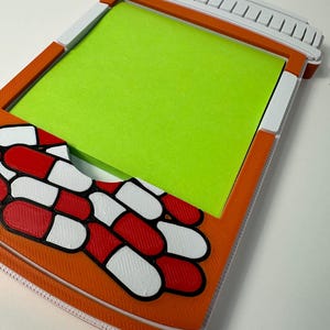 May include: An orange and white pill-shaped notepad holder with a green notepad inside. The holder is decorated with red, white, and black pill graphics. The top of the holder is white with orange accents.