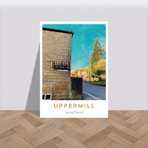 Puede incluir: Una impresión enmarcada que presenta una escena callejera en Uppermill, con un edificio de piedra y un letrero con el texto "Turog House". La impresión también incluye el texto "UPPERMILL" y "Turog House" en una fuente serif.