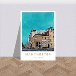 Puede incluir: Una impresión enmarcada con una fotografía de un edificio en Mánchester, Inglaterra, con el texto "MANCHESTER Crown & Kettle". El cielo es de color turquesa brillante.