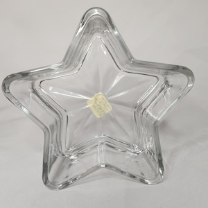May include: Clear glass star-shaped dish. The dish has a multi-pointed star design with a slightly raised rim. A small, rectangular sticker is affixed to the center of the dish. The dish is made of clear glass.