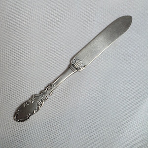 May include: A silver butter knife with an ornate handle and a rounded blade. The handle features detailed floral and scrollwork designs. The knife is set against a light grey background.