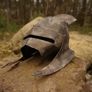 Peut inclure: Un casque en métal vieilli avec une visière et une crête pointue. Le casque est posé sur un rocher dans un environnement forestier.