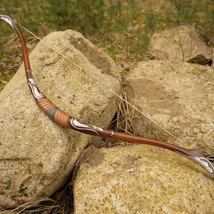 May include: A wooden bow with a brown and silver design. The bow is strung and resting on two large rocks.