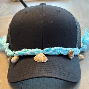May include: Black trucker hat with a mesh back and a curved brim. A light blue braided band is attached across the front, adorned with small seashells. The hat is a casual accessory.