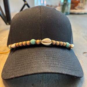 May include: Black trucker hat with a decorative band of wooden beads, turquoise beads, and a seashell. The hat has a curved brim and a mesh back.