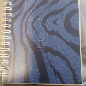 May include: A spiral-bound notebook with a blue and white zebra stripe pattern. The words "Atlya's Shop" are printed vertically on the cover. The notebook is open and lying on a gray surface.