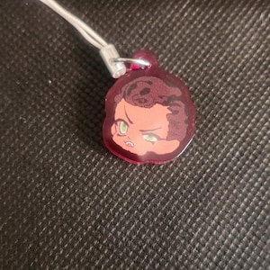 Puede incluir: Un pequeño dije acrílico de tono rojo que representa una cara estilizada con ojos verdes y cabello oscuro. Está unido a un cordón blanco con un cierre plateado. Probablemente un llavero o accesorio.