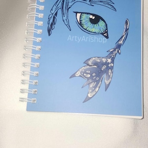 May include: Spiral-bound notebook with a light blue cover featuring an artistic eye and wing design in shades of blue. The text "ArtyArishop" is printed on the cover. The notebook is open, revealing the spiral binding.