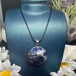 May include: A round pendant necklace with a dark blue and white stone set in a silver-colored metal frame. The pendant hangs from a black cord necklace. The necklace is displayed on a blue mannequin.
