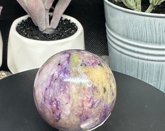 May include: A polished sphere with a mix of purple, yellow, and white hues. The gemstone ball is displayed on a black surface, with a US quarter coin in the foreground for size reference. Decorative plants are in the background.