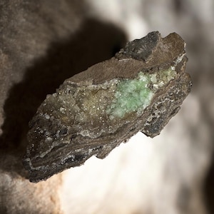 May include: A close-up of a rough-edged rock formation with a central pocket of light green crystals. The rock has layers of brown and gray tones, with a textured surface and a natural, earthy appearance.