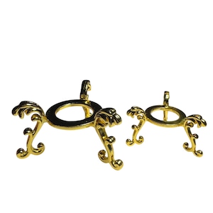 May include: Two ornate, gold-colored metal stands. Each stand features a circular ring with decorative, swirling accents and three curved legs. The stands are designed to hold a round object, such as a sphere or a decorative ball.