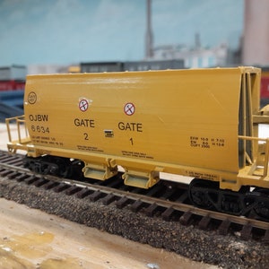 Puede incluir: Un vagón tolva modelo de ferrocarril amarillo con ruedas negras y una escalera de metal negro. El vagón está etiquetado como "OJBW 6834" y tiene dos puertas etiquetadas como "GATE 2" y "GATE 1". El vagón también tiene la siguiente información: "EXW 10-8 H 7-10 EW 8.0 H 112.8 CUFT 2300".