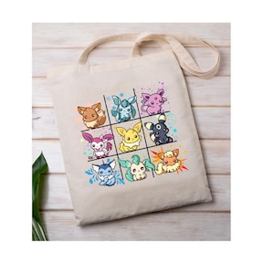 May include: A beige tote bag featuring a grid of nine colorful cartoon creatures. Each creature has unique features and colors, including brown, blue, pink, yellow, and black. The bag has a long strap for carrying and is made of a light-colored fabric.