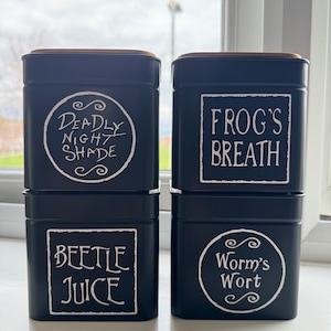 May include: Four black metal canisters with gold lids. The canisters are labelled with white text: "Deadly Night Shade", "Frog's Breath", "Beetle Juice", and "Worm's Wort".
