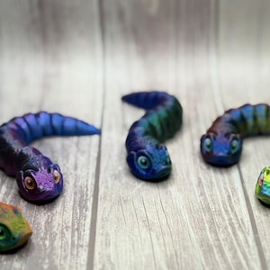 May include: A collection of five colorful, stylized snake figurines. Each snake has a unique color scheme, including shades of blue, green, and orange. The figurines are posed on a light wood-grain surface.