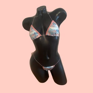 May include: A two-piece bikini set featuring a light blue fabric with a tropical palm tree and floral print. The triangle top and bottom pieces are trimmed with solid light pink fabric and connected by thin, delicate white string ties.