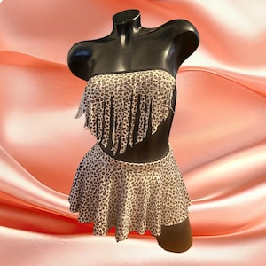 May include: A two-piece swimsuit set on a black mannequin. The set features a strapless top with fringe and a skirt, both in a leopard print pattern. The background is a soft, peach-colored fabric.