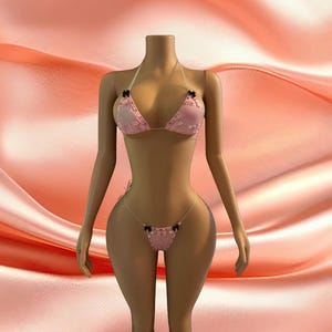 May include: A two-piece bikini set in light pink featuring triangle cups and a thong bottom. The set is adorned with small black bows at the strap connections and center of the bikini top and bottom, with clear plastic straps for support.