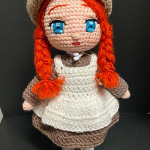 May include: A crocheted doll with red hair in braids, wearing a white apron over a brown dress and a brown hat. The doll has blue eyes and a pink face.